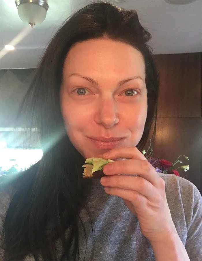 Celebrity without makeup, holding a sandwich in a casual home setting.
