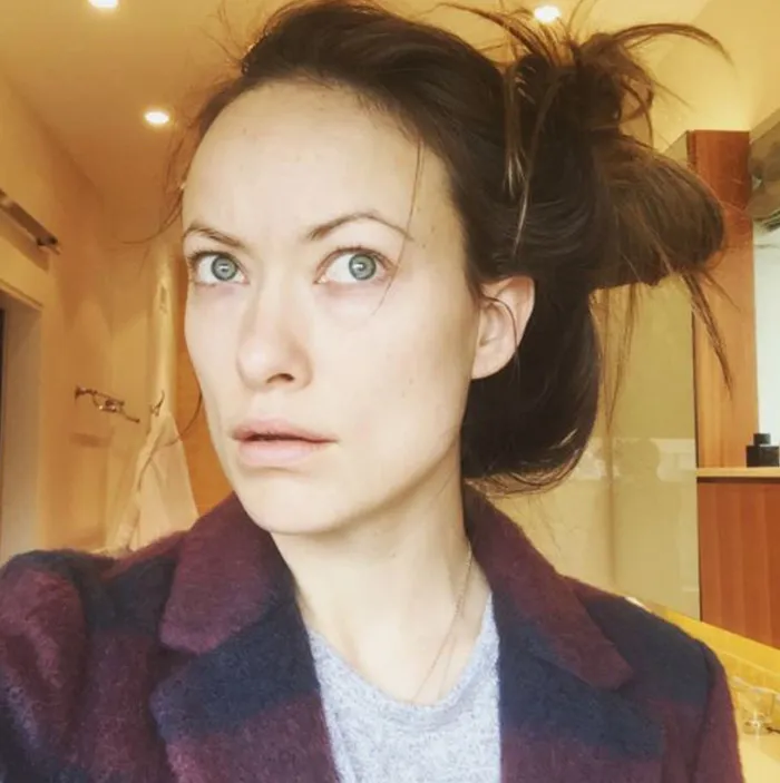 Celebrity without makeup, wearing a dark coat, hair up, standing in a warmly lit room.