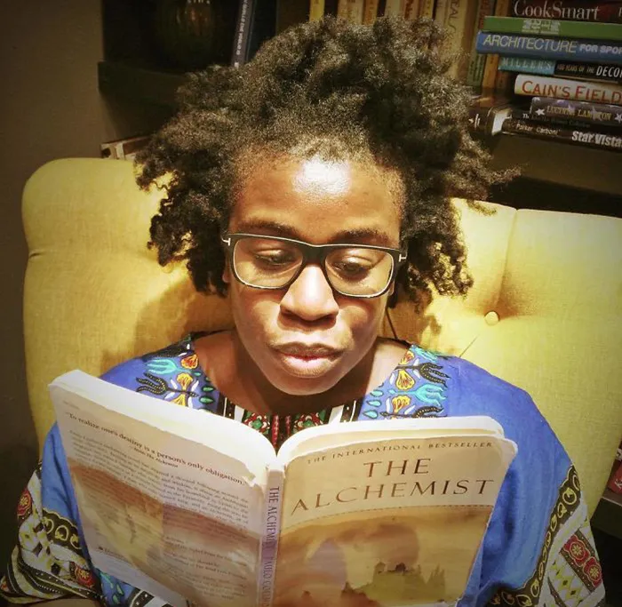 Person reading "The Alchemist" on a cozy chair, makeup-free, with natural hair and glasses, surrounded by books.