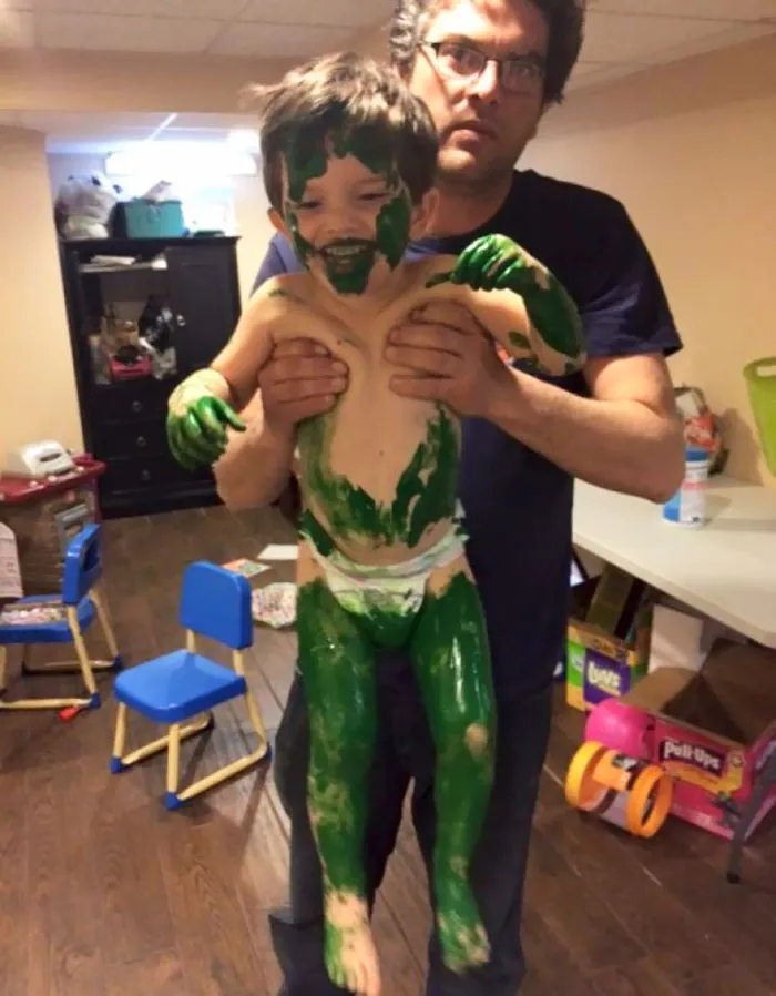 Child covered in green paint, held by an adult, illustrating a humorous moment when leaving kids alone.