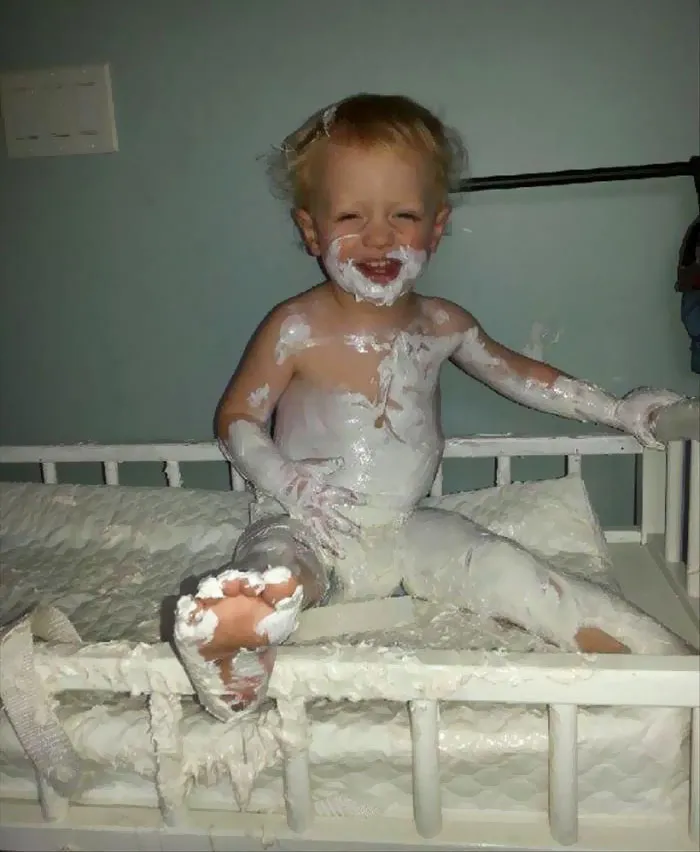 Child covered in cream and smiling, sitting on a bed, showing what happens when you leave kids alone.