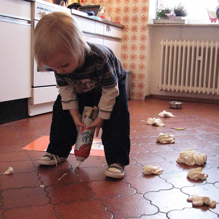 Child playing alone in kitchen, squeezing whipped cream onto the floor, showcasing a humorous and messy scene.