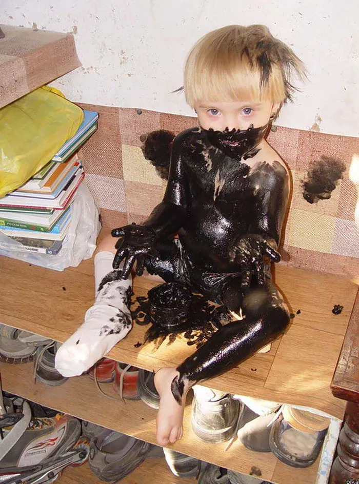 Child covered in black paint sitting on a wooden shelf, surrounded by shoes and books.