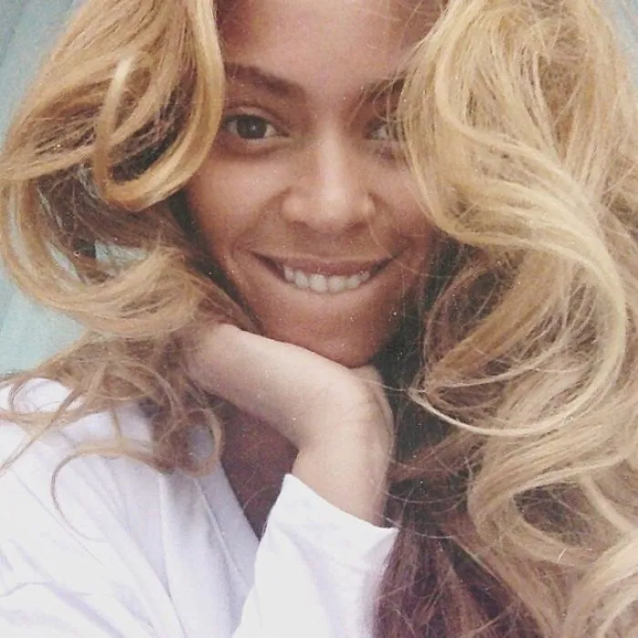 Celebrity posing without makeup, smiling with long wavy hair and a white shirt.