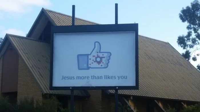 Funny church sign with a like button symbol reads, "Jesus more than likes you," outside a church building.