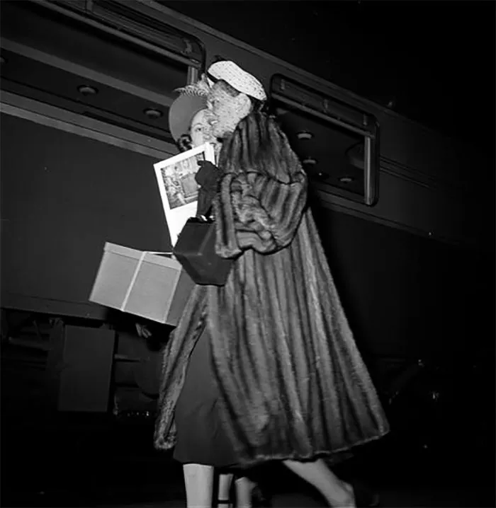 1940s New York photo by Stanley Kubrick, showing a woman in a fur coat, holding a package and newspaper at a train station.