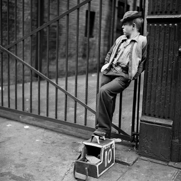 Teen boy in 1940s New York, captured by Stanley Kubrick, leaning on a gate with a shoe-shine box.