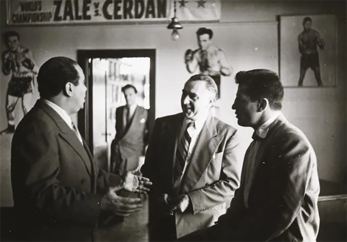 Three men in suits conversing in a 1940s New York boxing gym, with posters on the wall, photographed by Stanley Kubrick.
