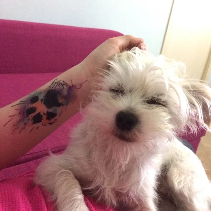 A person with a dog paw print tattoo on their arm rests next to a fluffy white dog on a pink couch.