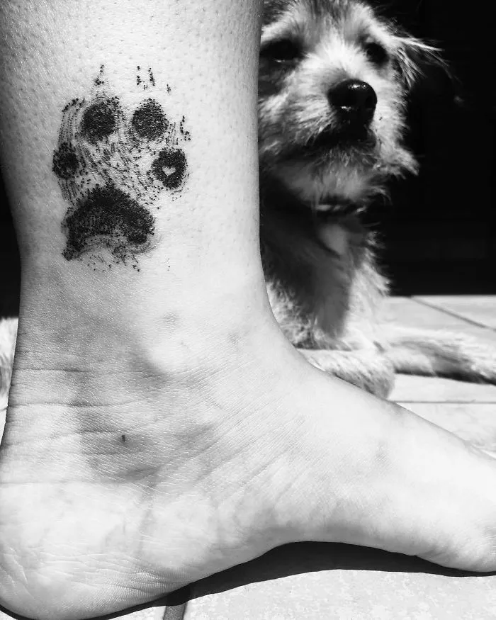 Dog paw print tattoo on a person's ankle with a furry dog in the background.