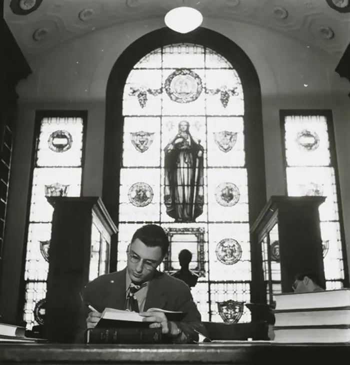 Young man in a suit and glasses, writing in a book at a library in 1940s New York.