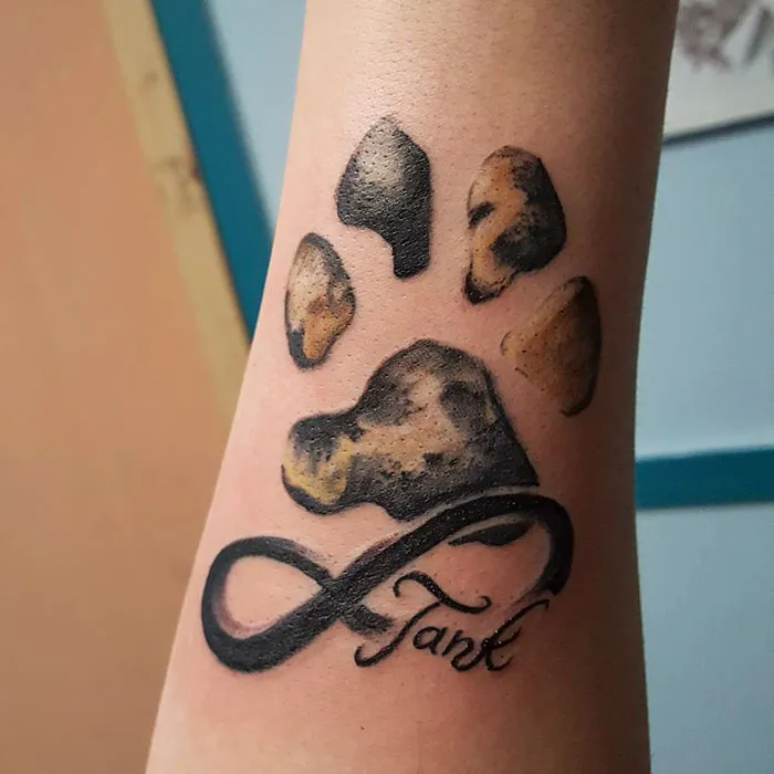 Tattoo of dog paw prints with an infinity symbol and the name "Tank" on a forearm.