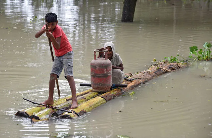 A Boy Rows A Makeshift Raft As He Transports A Woman And A Cooking Gas Cylinder Through The Flood Waters In Assam, India