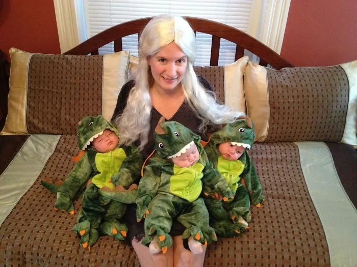 A mother dressed as Daenerys Targaryen sitting on a bed with her baby triplets in dinosaur Halloween costumes.