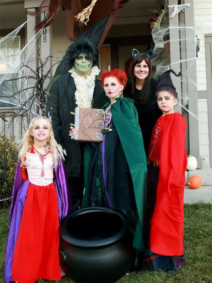 Family in Hocus Pocus themed Halloween costumes with a black cauldron in front of their decorated house.