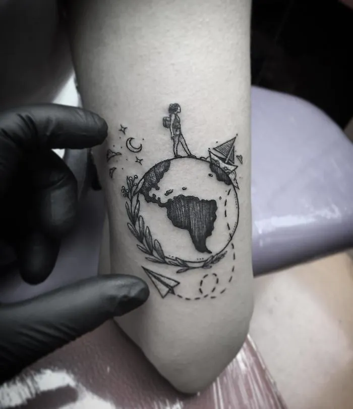 Travel tattoo featuring a traveler on a globe with paper planes and stars, inspiring adventure and wanderlust.