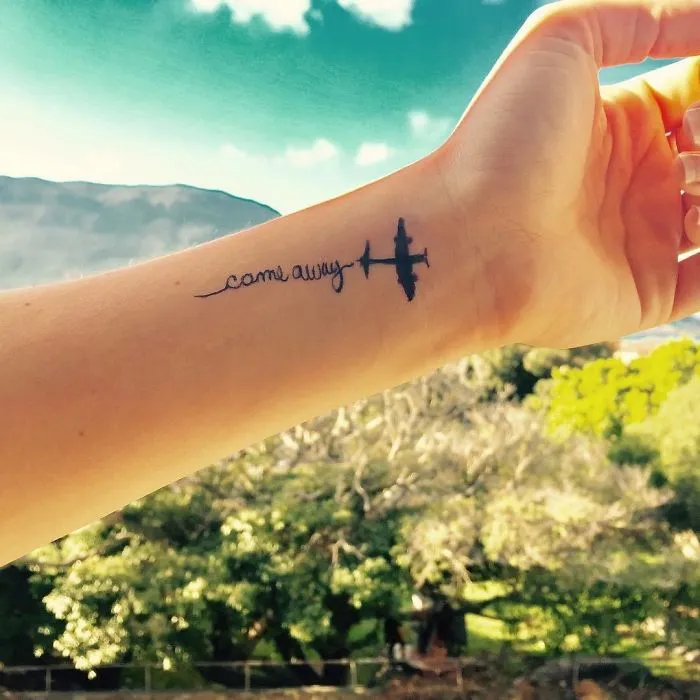 Travel tattoo featuring an airplane and "come away" text on a wrist against a scenic background.