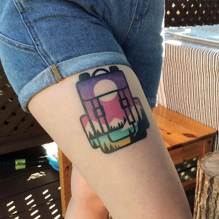 Colorful travel tattoo of a backpack with a sunset and mountains design on a person's thigh.