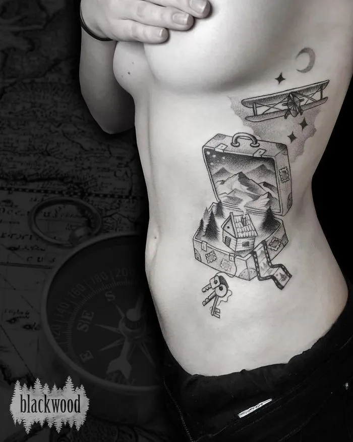Person with travel-themed tattoo featuring a suitcase, mountains, and a plane, symbolizing wanderlust.