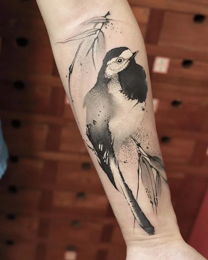 Watercolor tattoo of a bird on a person's forearm, featuring delicate black and gray ink strokes.