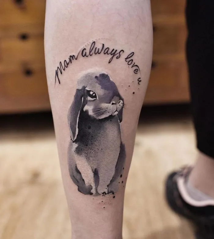 Watercolor tattoo by Chen Jie featuring a rabbit and the words "Mom always love u" on a person's calf.