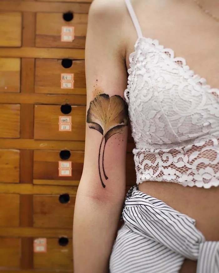 Watercolor tattoo of a ginkgo leaf by Chen Jie on a woman's arm, complementing her lace top and striped pants.