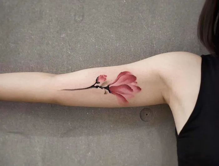 Watercolor tattoo of a pink flower on an arm, designed by Chen Jie, showcasing elegant artistry.