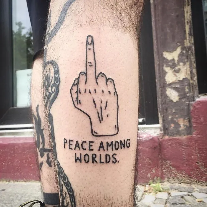Irreverent tattoo by German artist featuring a hand gesture with "Peace Among Worlds" text.