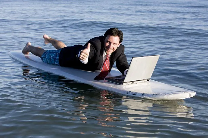 A happy man surfboarding in the water working on a laptop and showing thumb-up