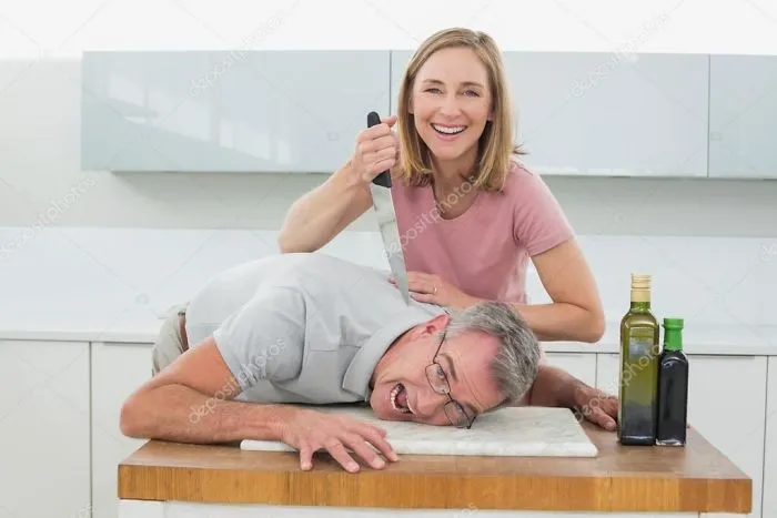 A happy woman holding a knife directed at an older man in the kitchen
