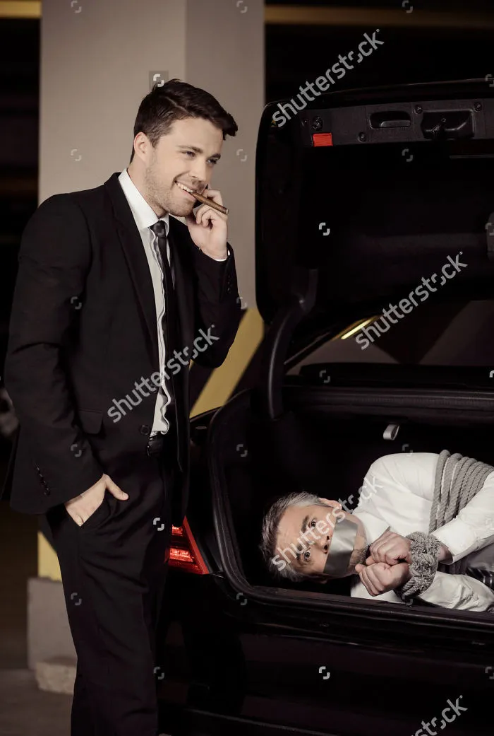 A man is casually smiling while taking a smoke break and talking on the phone, and there is a man tied up in his trunk