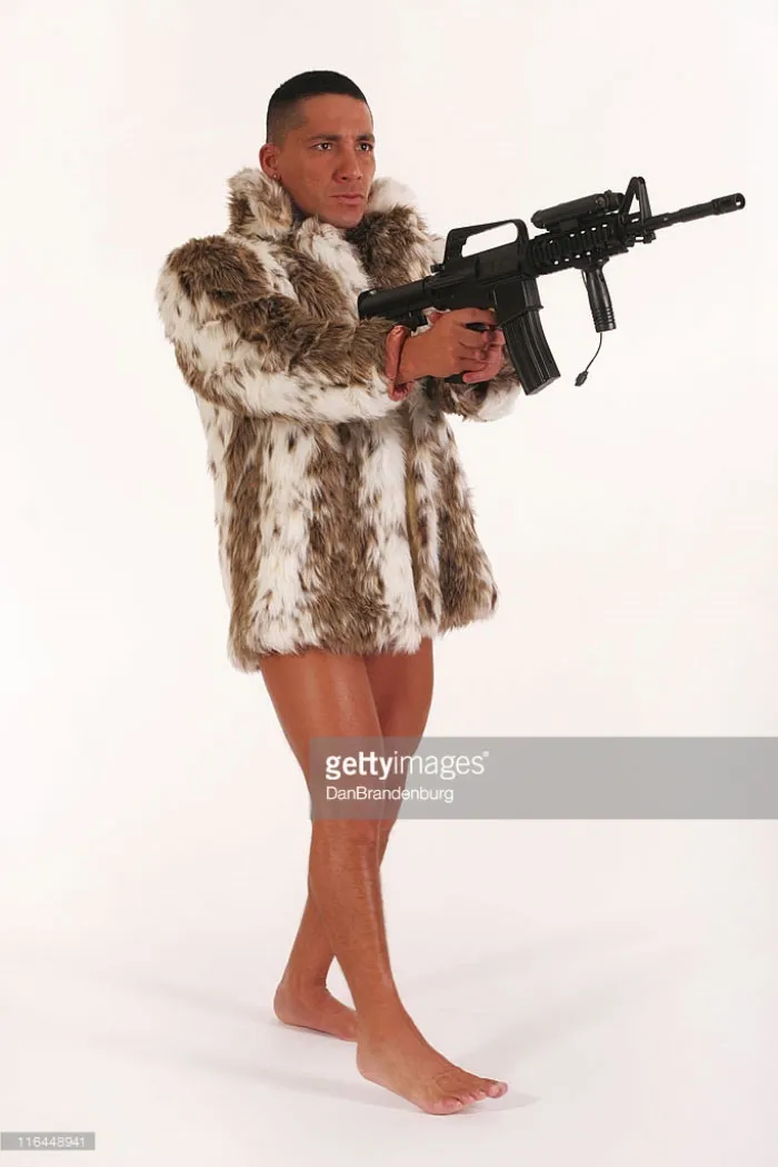 Naked man wears a fur coat and holds the gun in his hands