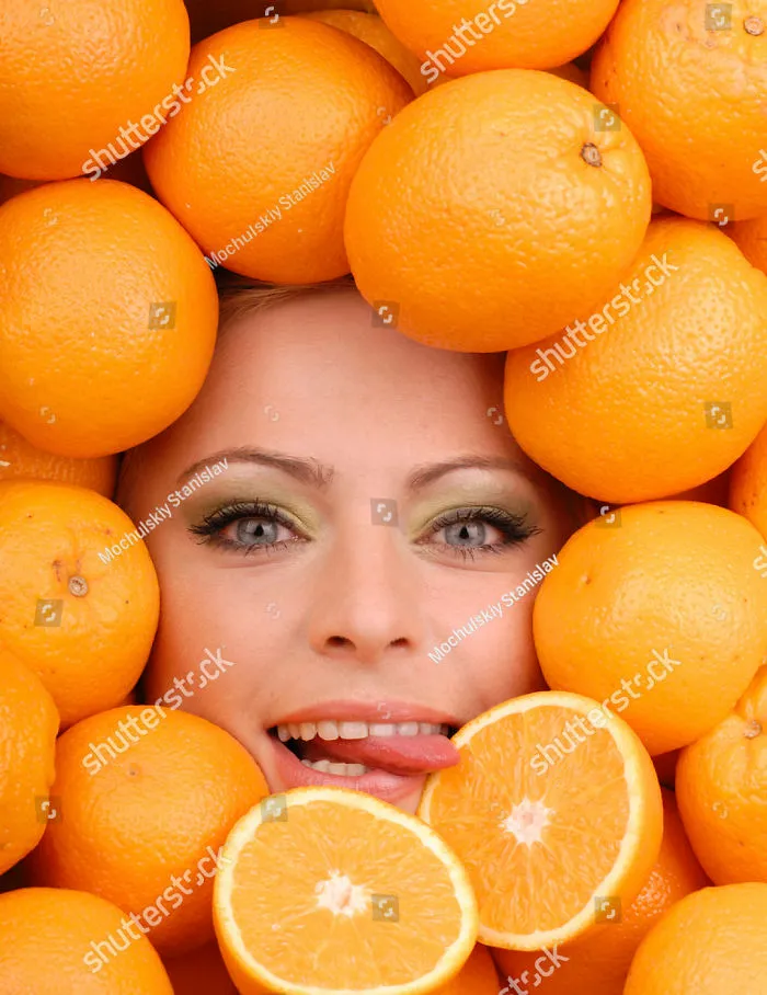 A woman's face among fresh oranges and she is licking one orange that is cut in half