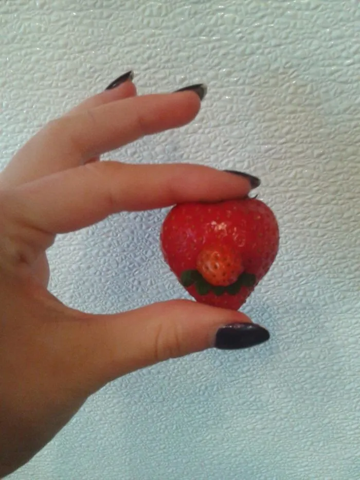 A hand holding a heart-shaped strawberry with a small strawberry on top, resembling an accidental cosplay.