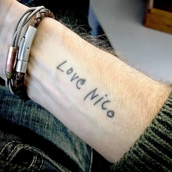 Tattoo with real meaning reading "Love Nico" on a forearm, beside a braided bracelet.