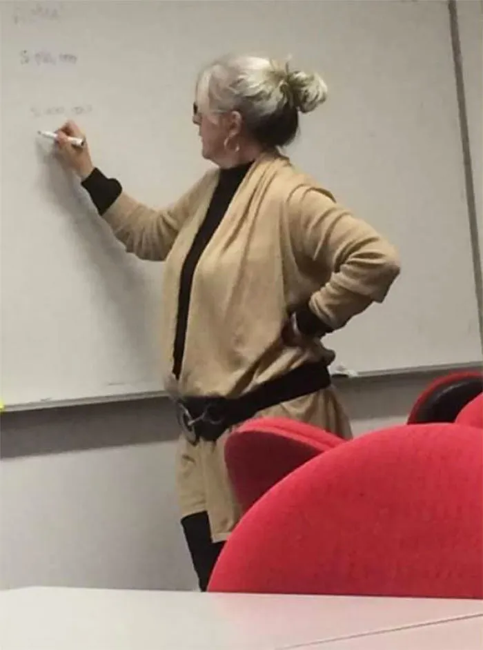 Woman in beige outfit unintentionally resembling a cosplay character, writing on a whiteboard in a classroom setting.