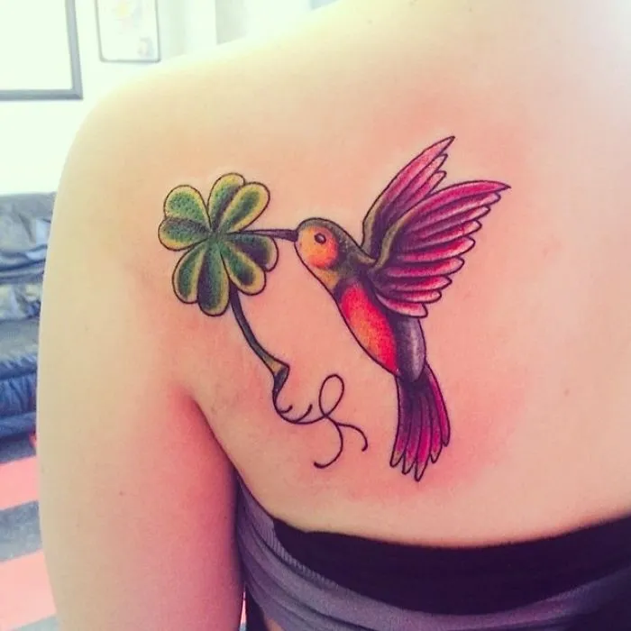 Colorful hummingbird tattoo holding a four-leaf clover on shoulder, symbolizing powerful stories and real meaning.