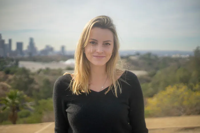 Young woman outdoors with a city skyline in the background, focused on interviewing people on their birthdays.
