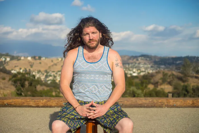 Man with long hair and tattoos sitting outdoors on a stool, part of interviewing 35 people on their birthdays series.