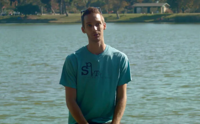 Man sitting by a lake, casually dressed, engaging in an informal conversation about interviewing people on birthdays.
