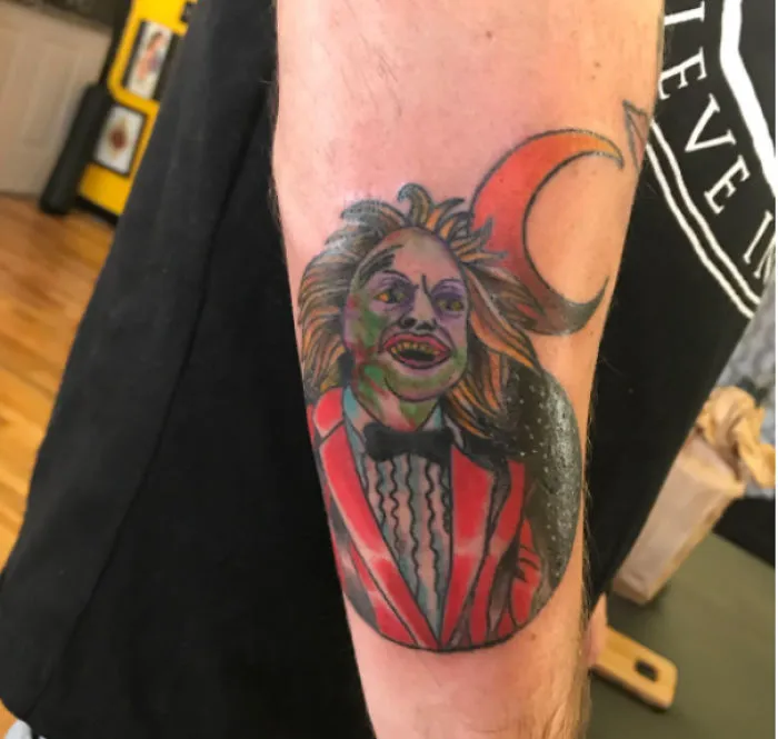 Bad tattoo fail of a colorful character in a red suit with a creepy grin and a crescent moon on an arm.