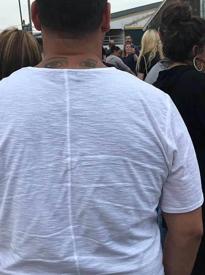 Man with eyes tattooed on the back of his neck, wearing a white shirt in a crowd. Tattoo fails humor.