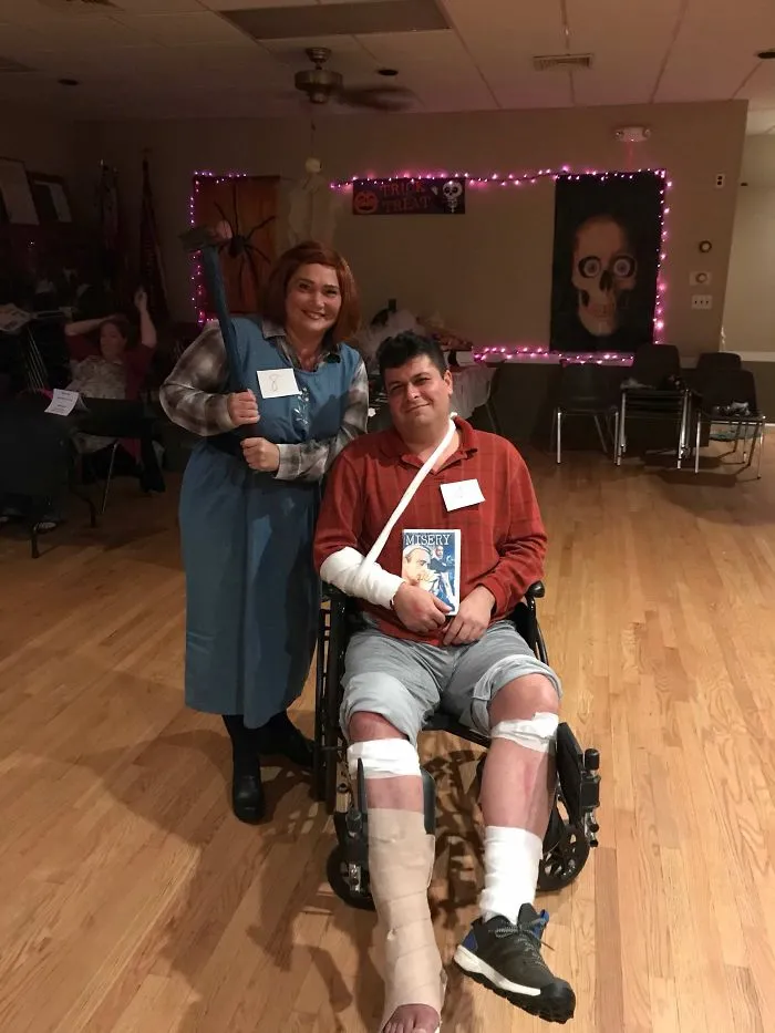 Couple dressed as characters from "Misery" for Halloween, one in a wheelchair with bandages, the other holding a fake hammer.
