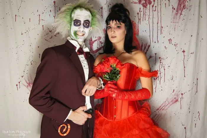 Couple in Halloween costumes, one in a red dress with roses, the other in a painted face and suit, posing against a spooky backdrop.