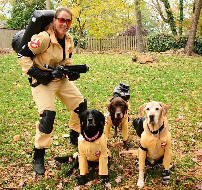 A woman and three dogs dressed up in matching Ghostbusters theme Halloween costumes outdoors.