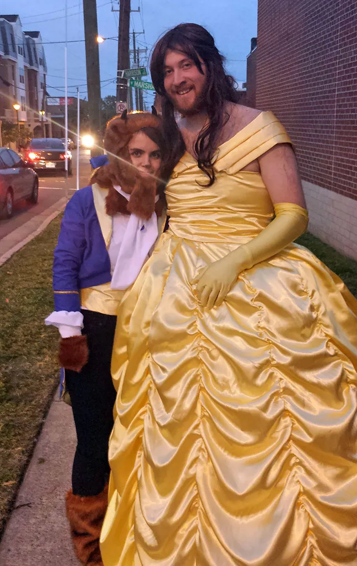Couple in creative Halloween costumes, with one dressed as a beast and the other in a yellow ball gown, smiling on the street.
