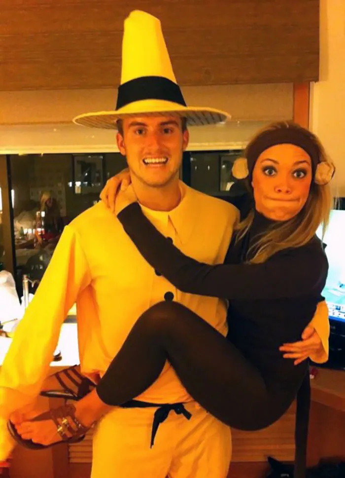 Couple in creative Halloween costumes; one as the Man in the Yellow Hat, the other as a monkey, showcasing Halloween spirit.
