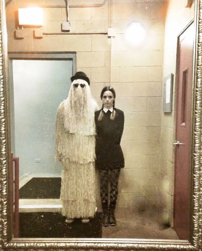 Couple in Addams Family costumes, reflecting in a mirror. Halloween outfits with creativity and style.