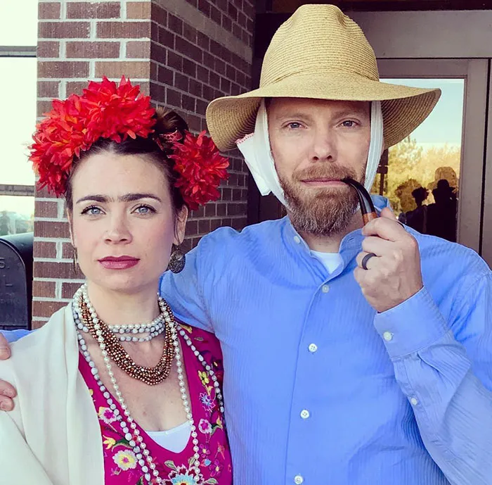 Couple dressed in creative Halloween costumes, with floral headpiece and straw hat, embodying artistic characters.