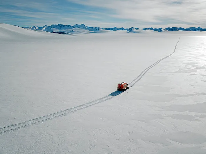 Snow vehicle traveling across vast icy landscape in Antarctica with distant mountain range under clear sky